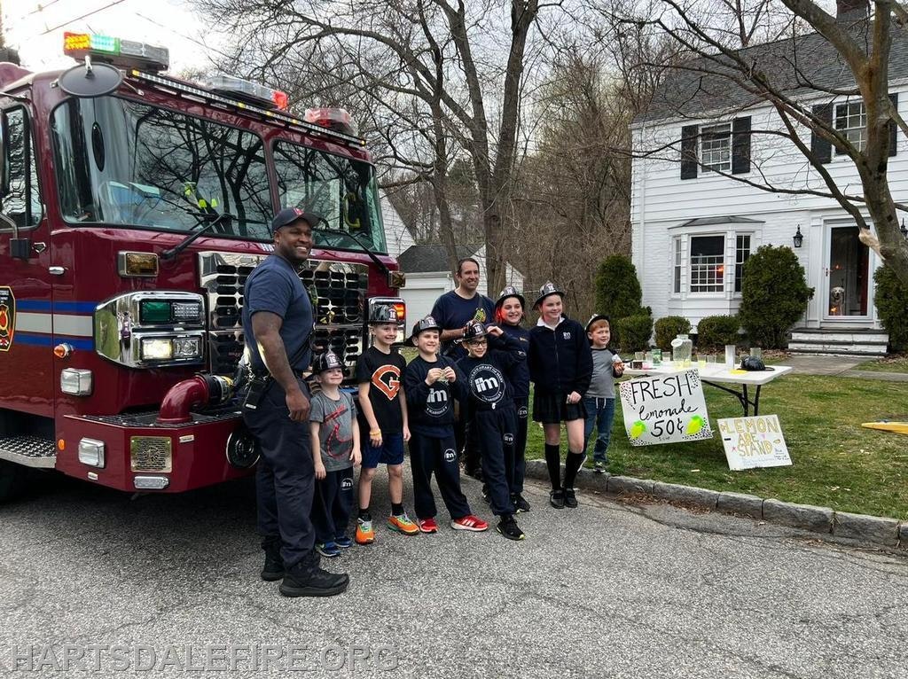 A group of kids and firefighters pose by a fire truck, promoting a lemonade stand with signs indicating prices.