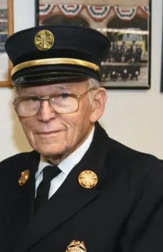 An elderly man in a formal uniform and hat, likely representing a fire or emergency service, posing for a photo.