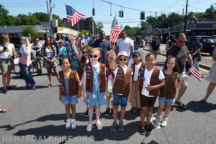 A group of children in brown vests, holding American flags, participating in a parade with adults in the background.