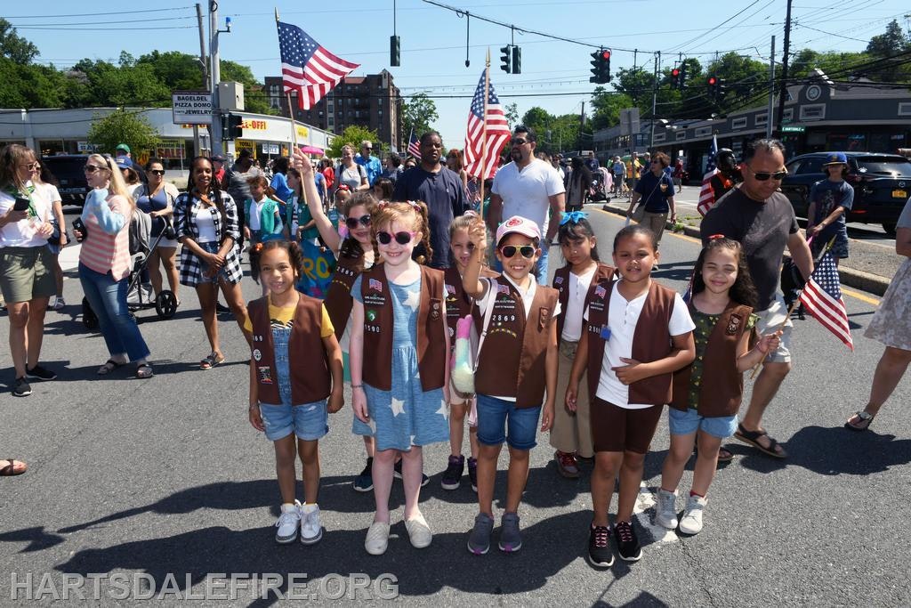 A group of children in brown vests, holding American flags, participating in a parade with adults in the background.