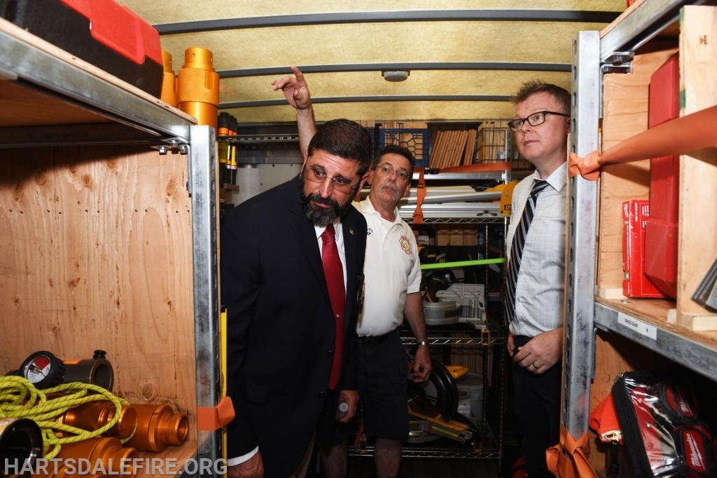 Three people examining equipment inside a storage area or trailer, with shelves holding various tools and binders.