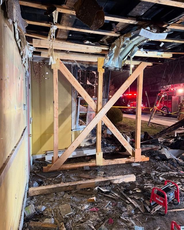 The image shows a damaged interior of a building with wooden support beams, debris on the floor, and emergency vehicles outside.