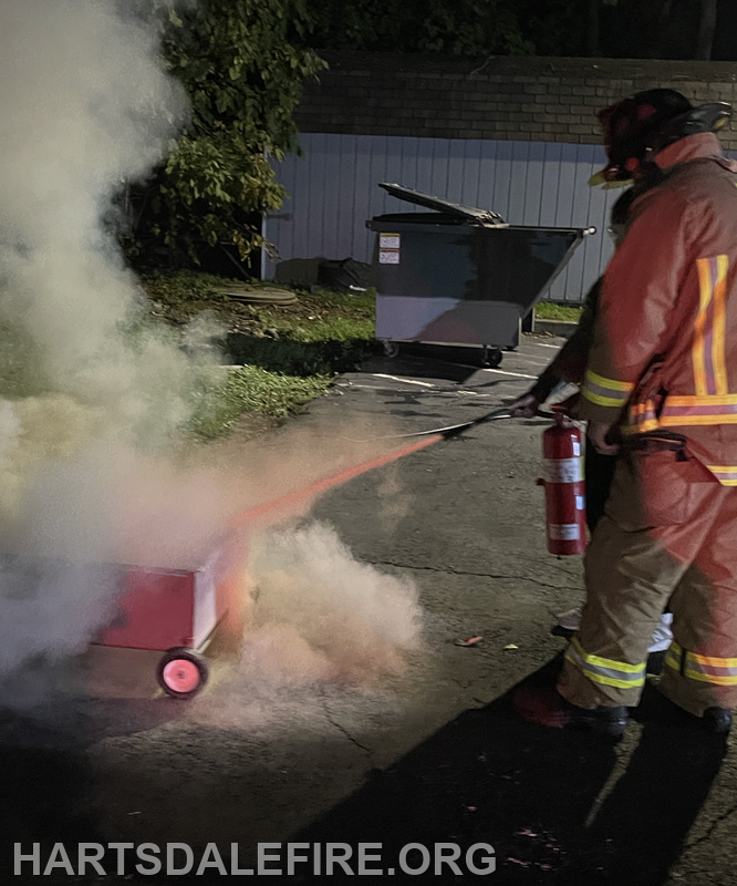A firefighter is using a fire extinguisher to put out flames from a small cart, creating smoke in a night setting.
