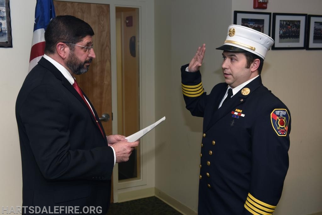 A formal ceremony with a man in a suit holding a document and a uniformed firefighter raising their right hand, next to a flag.