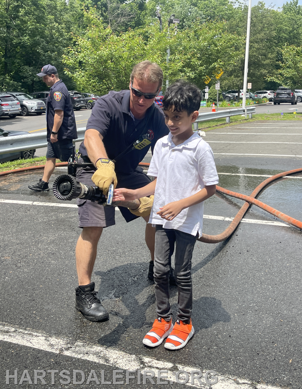 A firefighter teaches a young boy to handle a fire hose, both smiling in a parking area with fire equipment around.
