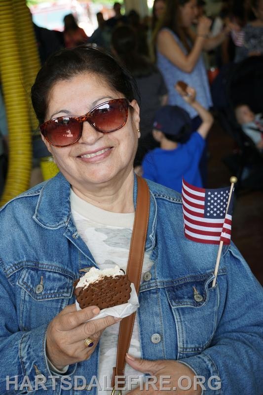 A person wearing sunglasses and a denim jacket holds an ice cream sandwich and a small American flag.