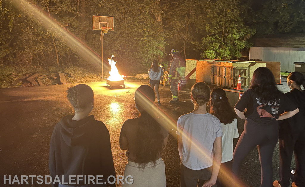 A group watches a firefighter manage a controlled fire outdoors near a basketball hoop, with safety cones and structures around.