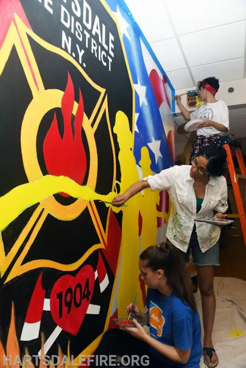 Three people painting a vibrant mural for "Hartsdale Fire District N.Y." with fire imagery and stars.