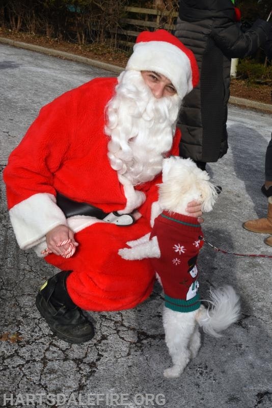 Santa kneeling with a small white dog wearing a festive sweater, holding a candy cane.