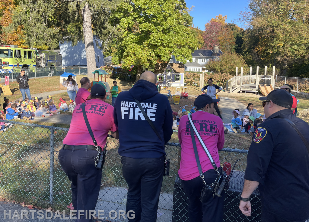Firefighters watch as children gather outside for an event, with playground equipment and a fire truck in the background.