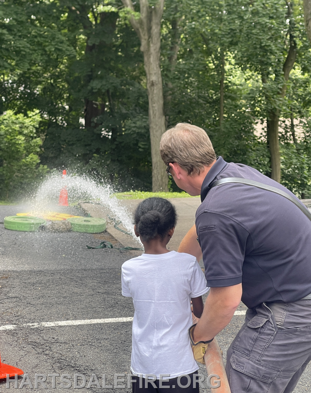 A child learns to use a fire hose under the guidance of an adult, with water spraying out in a lush green setting.