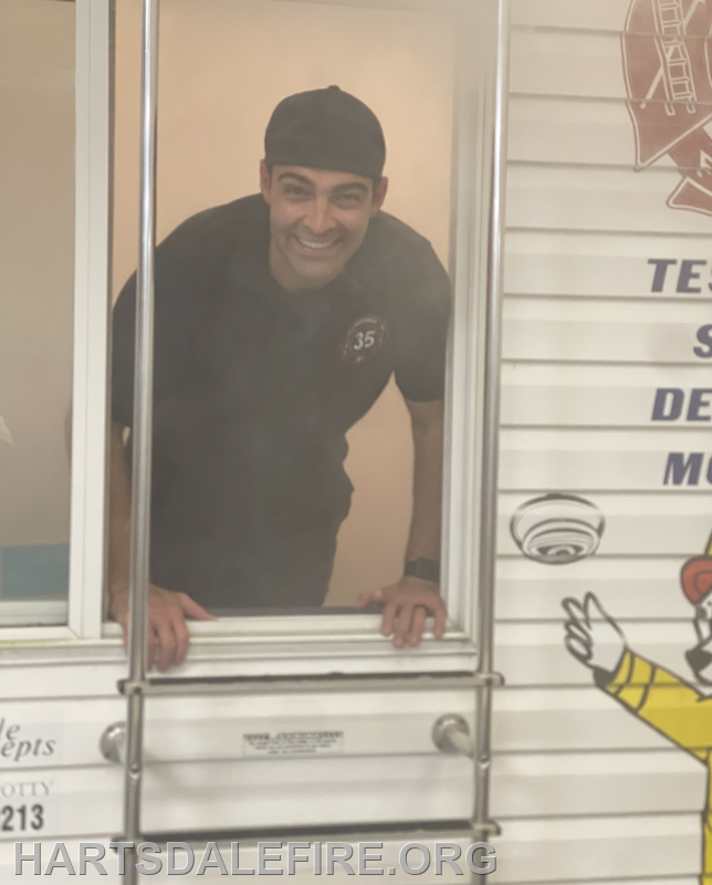 A smiling person in a black shirt is leaning out of a window, with a fire department logo visible in the background.
