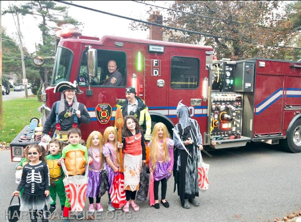 A fire truck is parked, and children in Halloween costumes are posing in front of it, holding trick-or-treat bags.