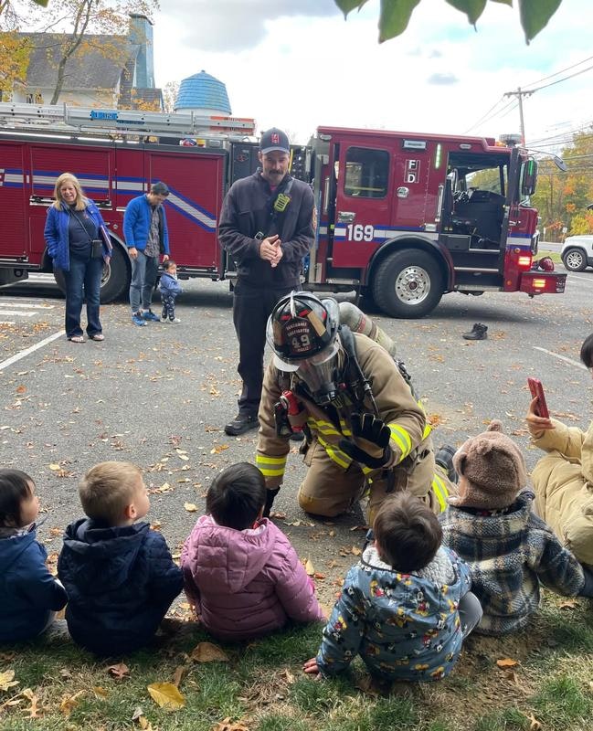 A firefighter engages with children sitting on the ground, while a fire truck is parked nearby and adults watch.