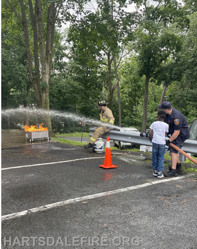 A firefighter sprays water on a controlled fire while a child and another officer assist in a training exercise.