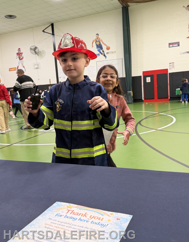 A child in a firefighter costume holds a walkie-talkie while a girl smiles behind him in a gym setting.