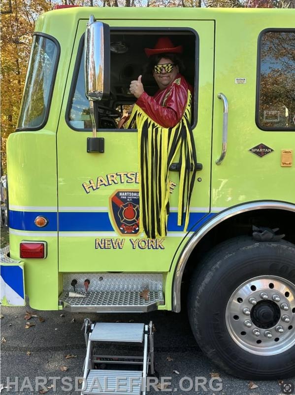 A person in a vibrant costume with a red hat and sunglasses gives a thumbs up from inside a green fire truck in Hartsdale, NY.