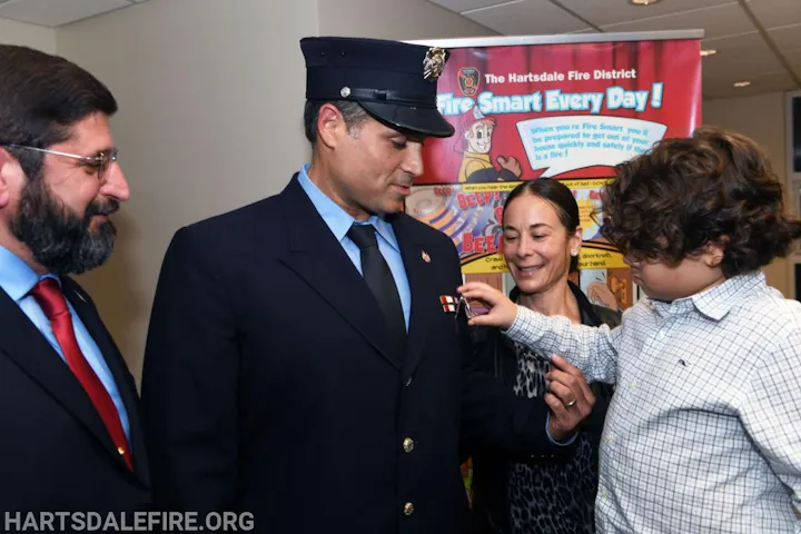 A firefighter receives a pin from a child, with a woman and another man nearby, against a fire safety poster.