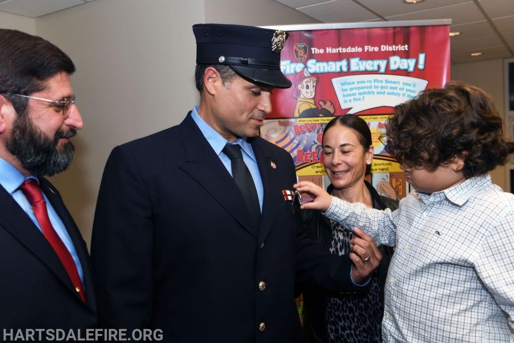 A firefighter receives a pin from a child, with a woman and another man nearby, against a fire safety poster.
