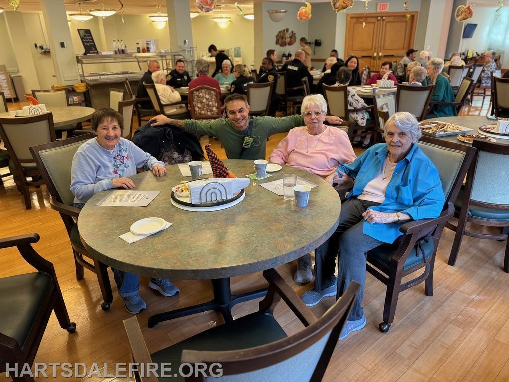 A group of four people smiles around a table in a dining area, while others socialize in the background.