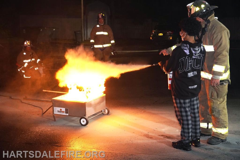 A firefighter guides a person using a fire extinguisher on a controlled fire.