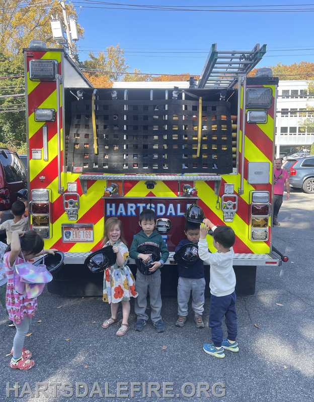 Children in front of a fire truck, holding black helmets and smiling. A vibrant fall backdrop completes the scene.