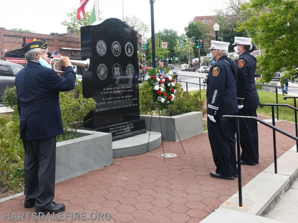 A solemn ceremony with three uniformed men in front of a memorial wall, one playing a bugle, honoring military service.