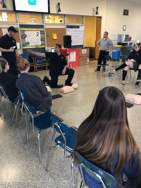 CPR training session with instructor demonstrating chest compressions on a dummy, participants seated and observing.