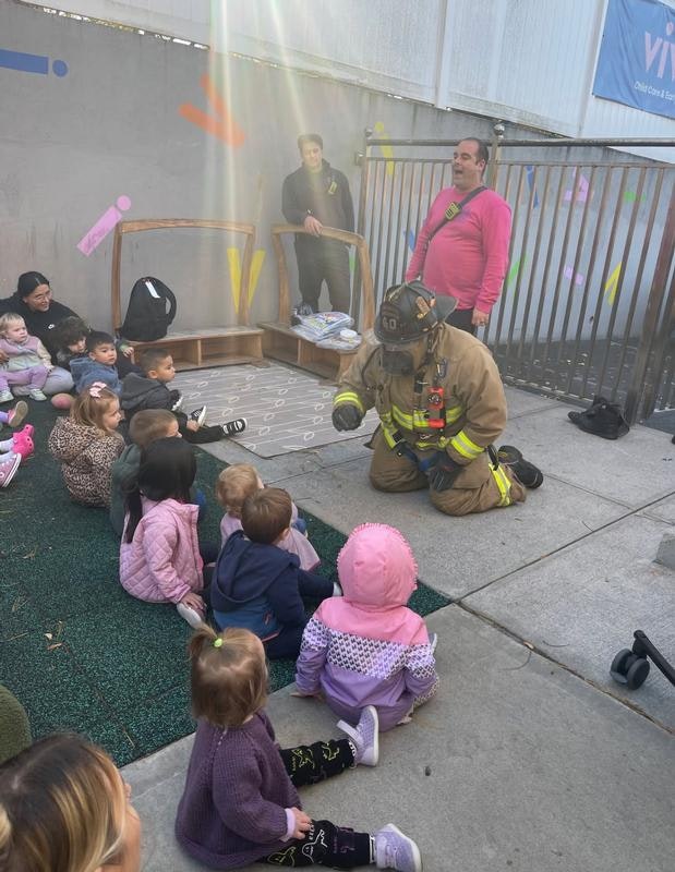 A firefighter engages with a group of young children sitting on the ground, while adults observe nearby.