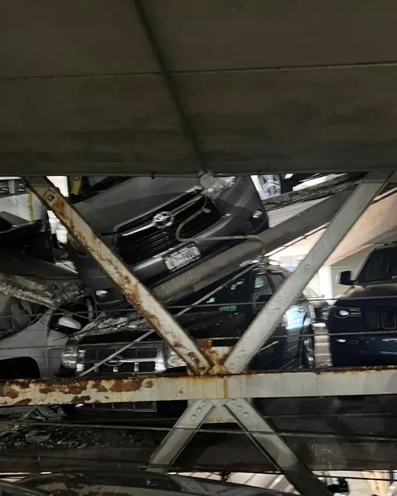 A chaotic scene in a parking garage with multiple cars piled up and partially collapsed structure visible among them.