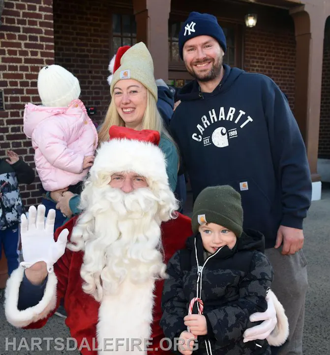 A family poses with Santa Claus, smiling and dressed warmly, in a festive outdoor setting.