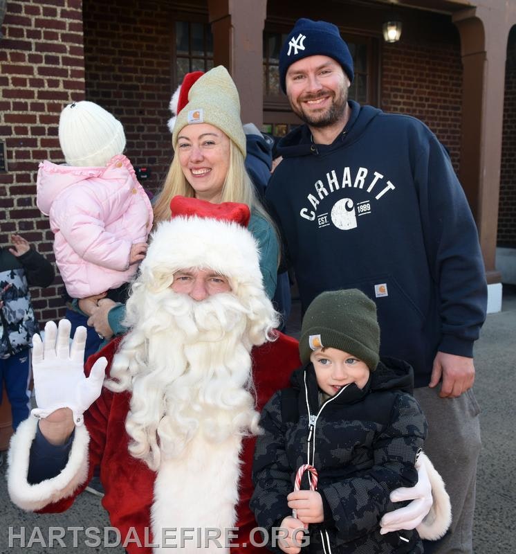 A family poses with Santa Claus, smiling and dressed warmly, in a festive outdoor setting.