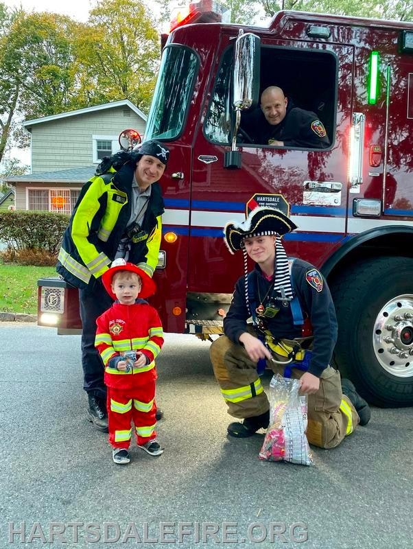 A young child in a firefighter costume poses with firefighters by a fire truck, creating a festive scene.