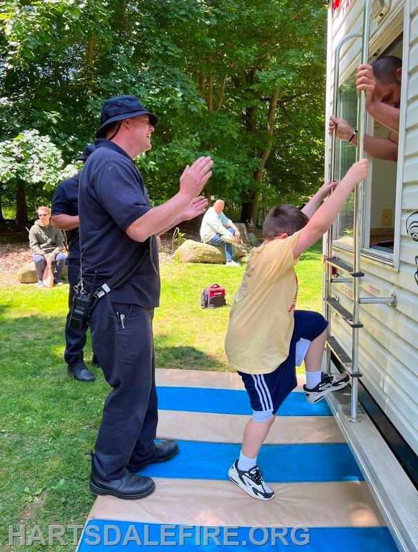 A child climbs into a trailer with assistance from a person outside, while another person watches. It's a sunny day in a park setting.