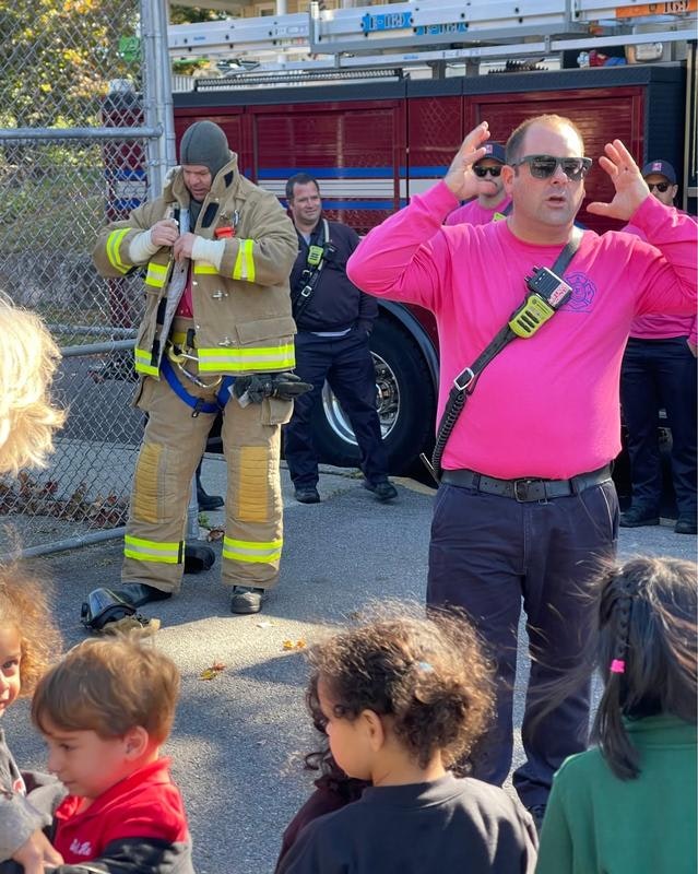 A fire safety event with firefighters, a crowd of children, and a firefighter in gear. Engaging presentation in progress.
