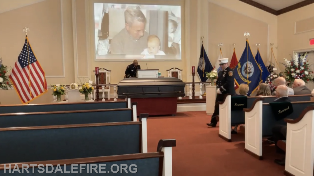 A memorial service with a casket, an image projected on the wall, and attendees, including a uniformed officer.
