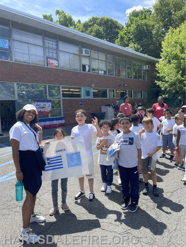 A group of children, some holding a sign for Greece, are outside a school during a sunny day, likely for an Olympic-themed event.