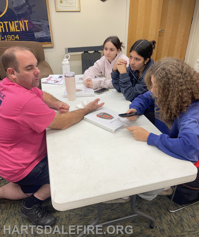 A man teaches a group of three girls at a table, engaged in discussion, with materials related to a fire department.