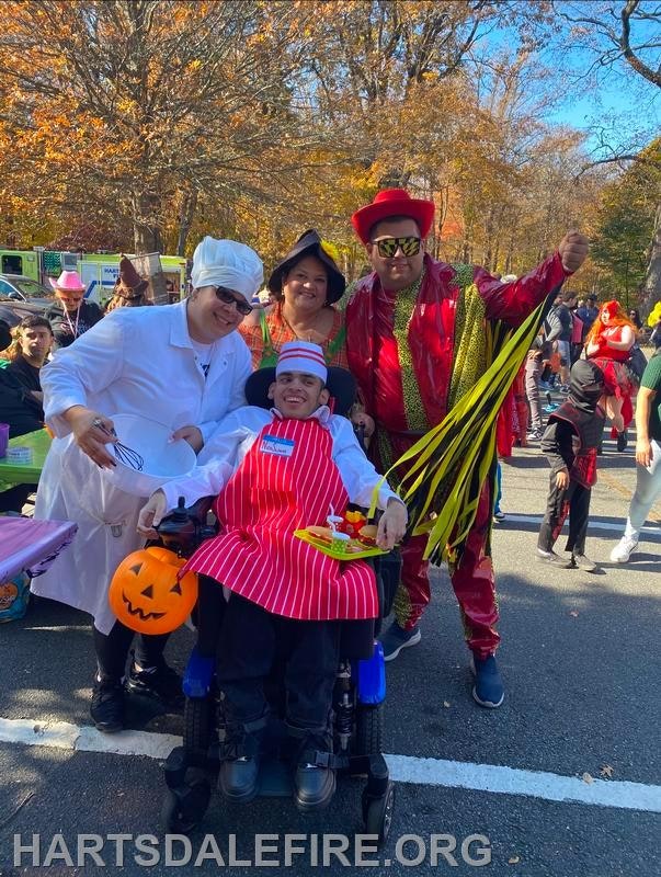 A festive group in costumes at an event, featuring a chef, a woman, and a man in shiny attire, with a child in a wheelchair.