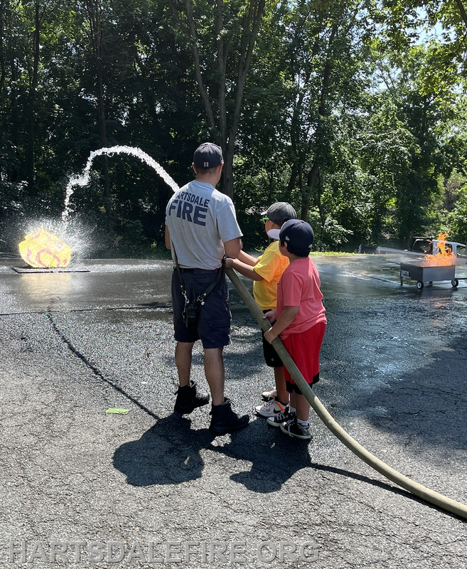 Firefighter and two kids practice using a hose to extinguish a small fire outdoors.