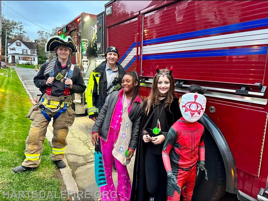 Firefighters with kids in costumes, including a pirate and superhero, by a fire truck during an event.