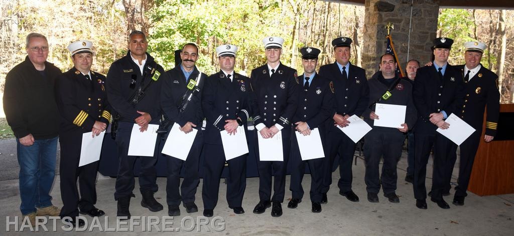 A group of firefighters and officials stands together, holding certificates in a park setting, celebrating achievements or awards.