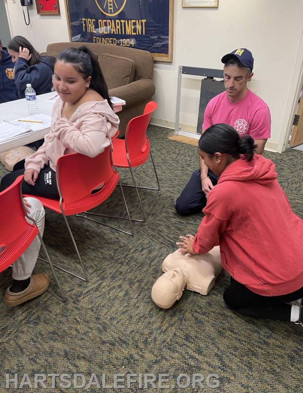 A group learning CPR, with one person practicing on a CPR dummy while others observe in a classroom setting.