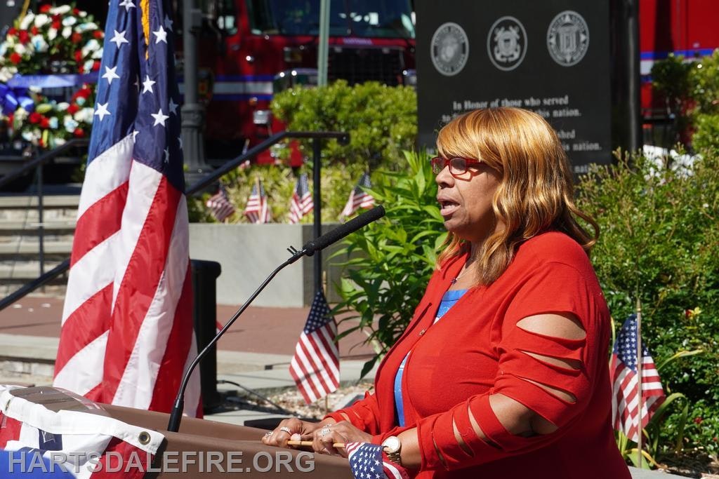 An event honoring service members features a speaker by a podium, with flags and a fire truck in the background.