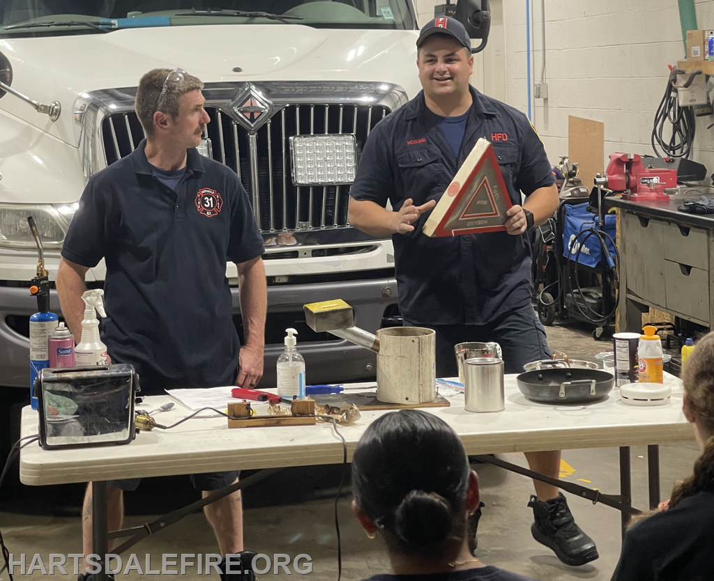 Two firefighters conduct a safety demonstration in a garage, showing tools and equipment to an attentive audience.
