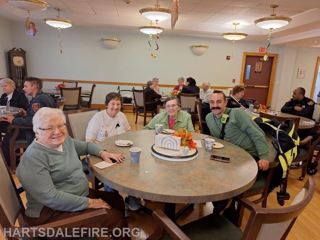 A cheerful group of seniors and a few staff members gather around a table, enjoying refreshments and conversation in a cozy setting.