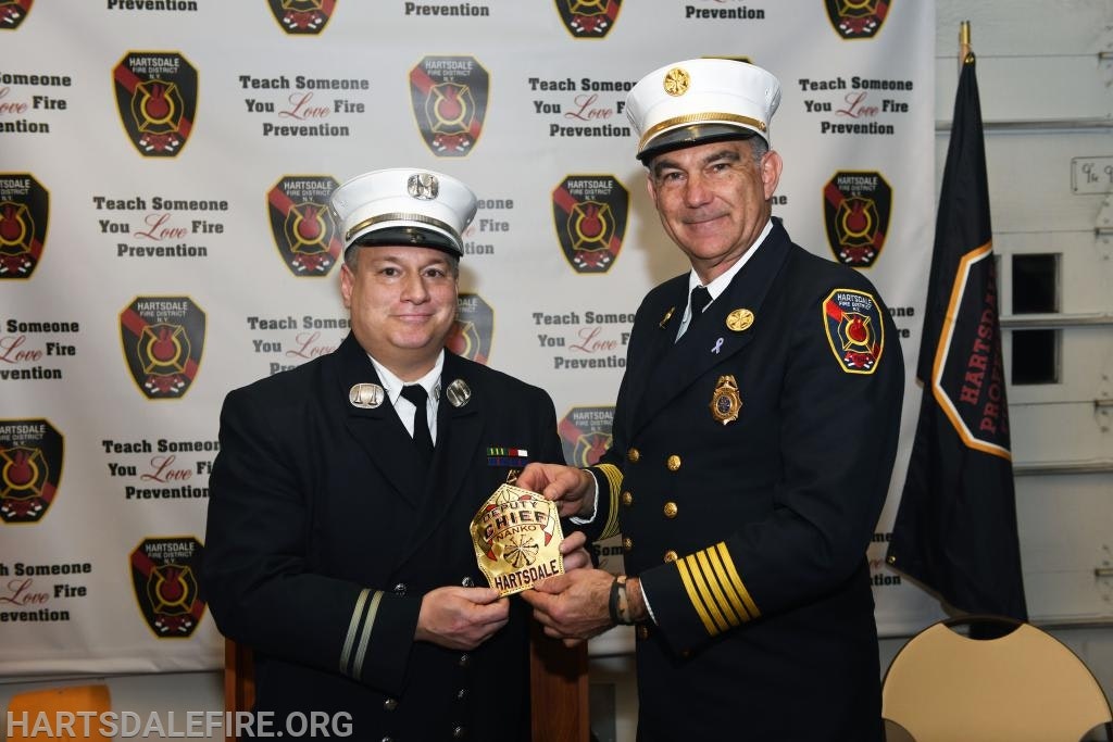 Two men in fire uniforms shaking hands, exchanging a badge. Background displays fire prevention messages and logos.