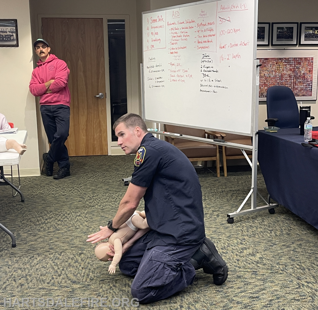 A firefighter demonstrates CPR techniques on a training mannequin in a classroom setting, while others observe.