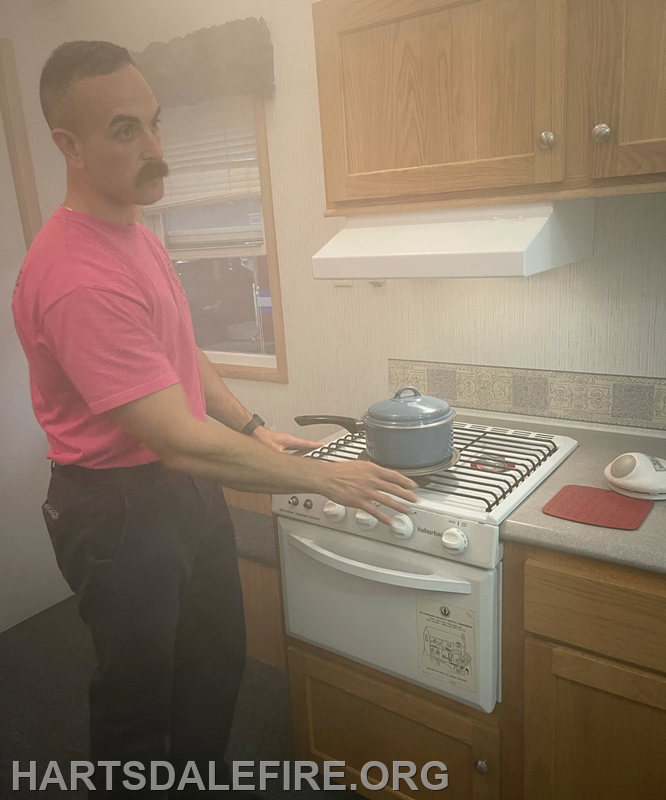 A person in a pink shirt is at a kitchen stove with a pot, surrounded by smoke, indicating a potential cooking hazard.