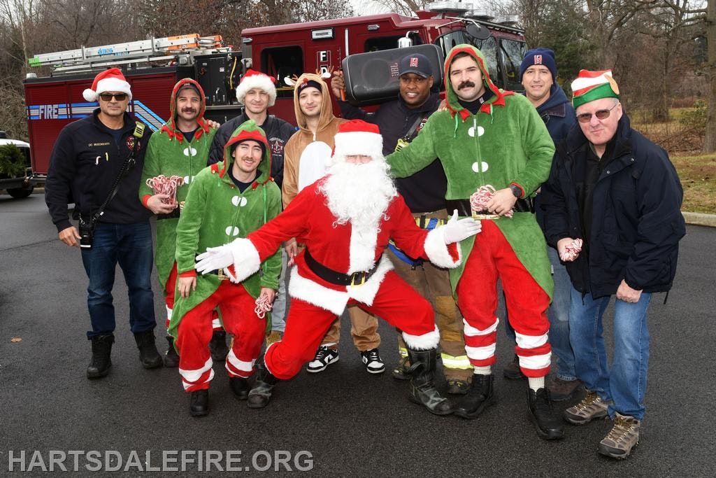 A festive group of people in holiday costumes, including Santa and elves, posing in front of a fire truck.